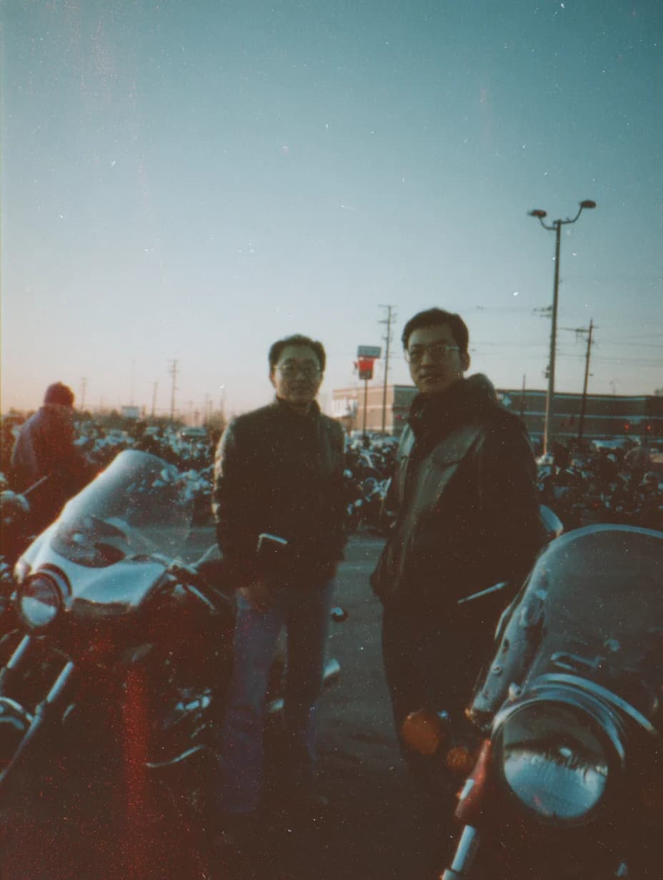 Two riders standing among motorcycles at dusk, in a faded early-2000s snapshot