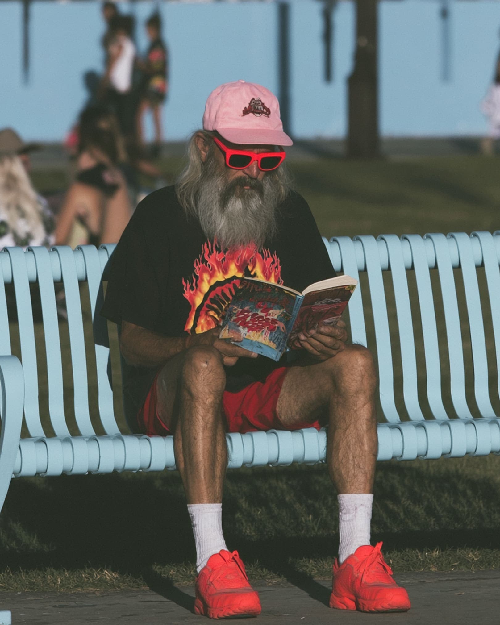 An older man with a long grey beard, a pink cap and red sunglasses reads a paperback on a sunlit boardwalk bench