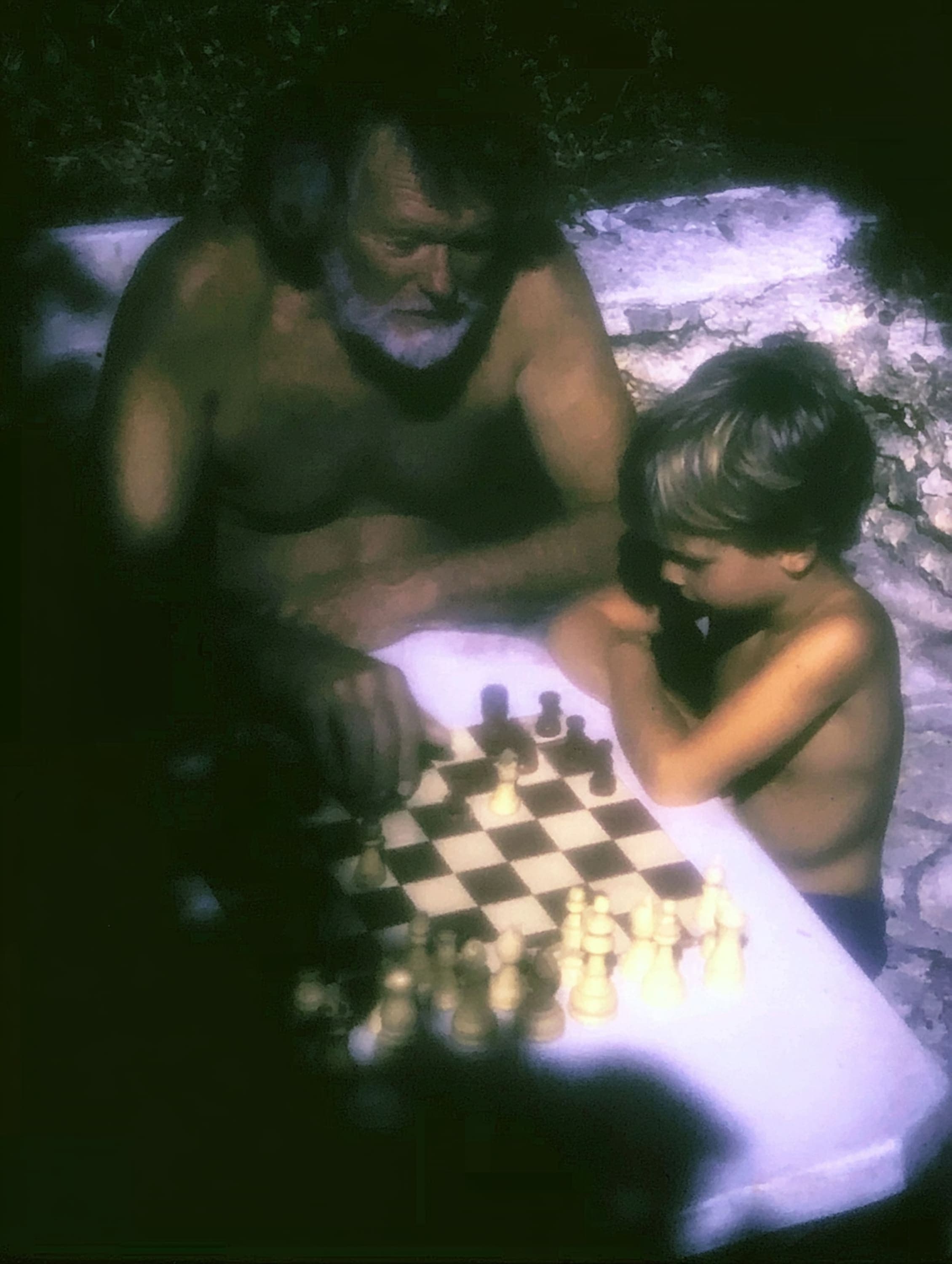 An older man and a young boy lean over a chess game on a white table in fading evening light