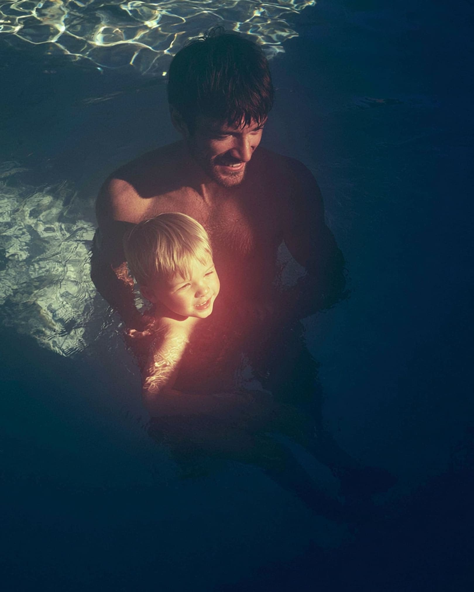 A man holds a laughing toddler in dark water, both lit from above
