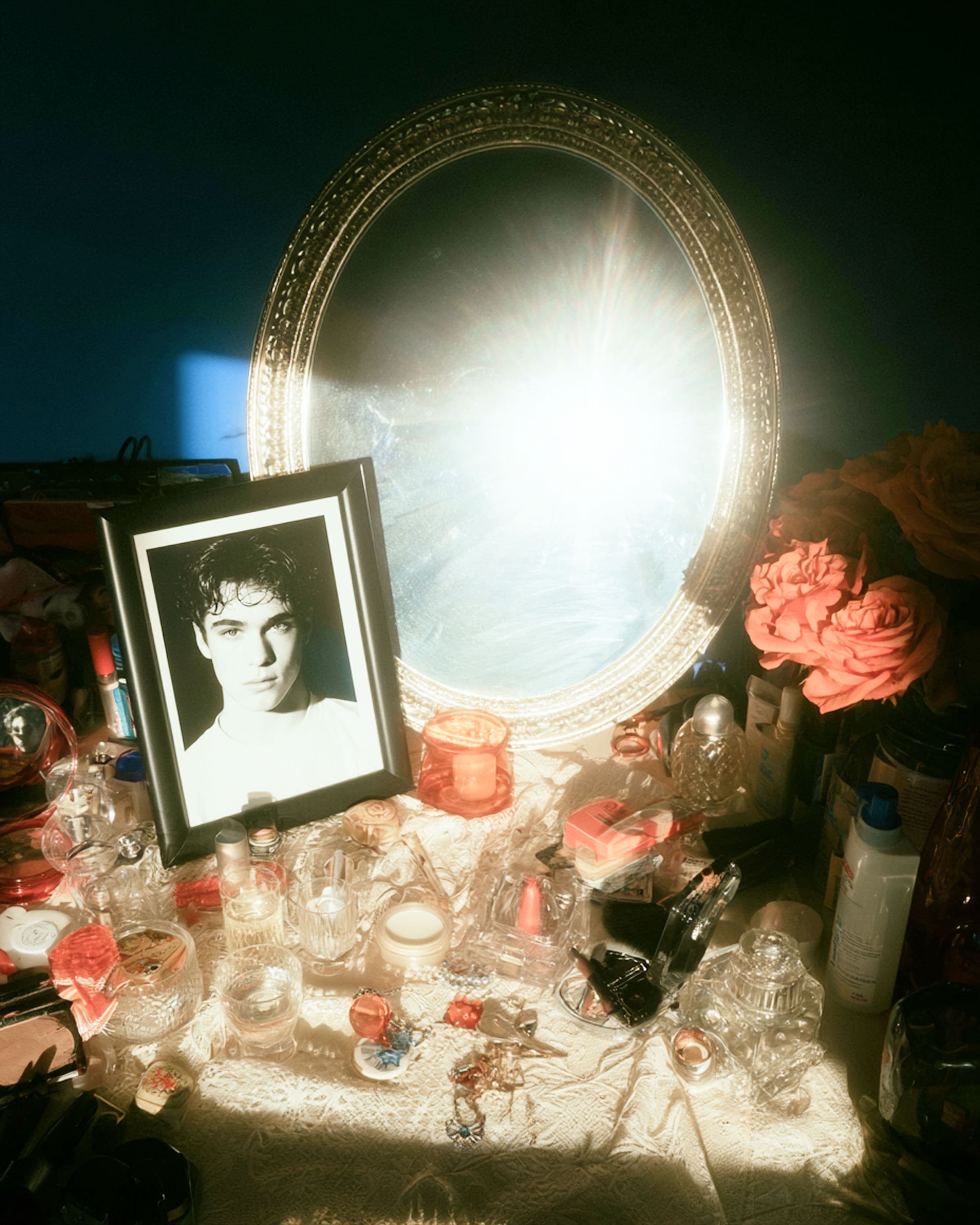 A framed black-and-white portrait of a young man stands among perfume bottles and roses on a dressing table, beside a brightly lit oval mirror