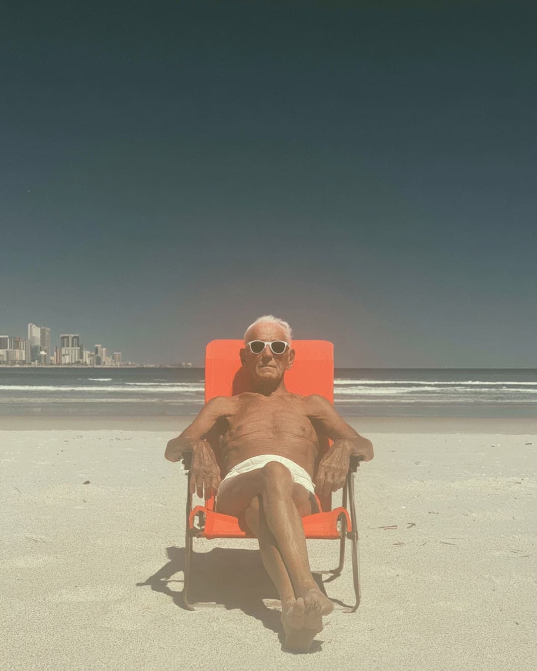 An older man reclines alone in an orange beach lounger on empty sand, a distant city skyline behind him