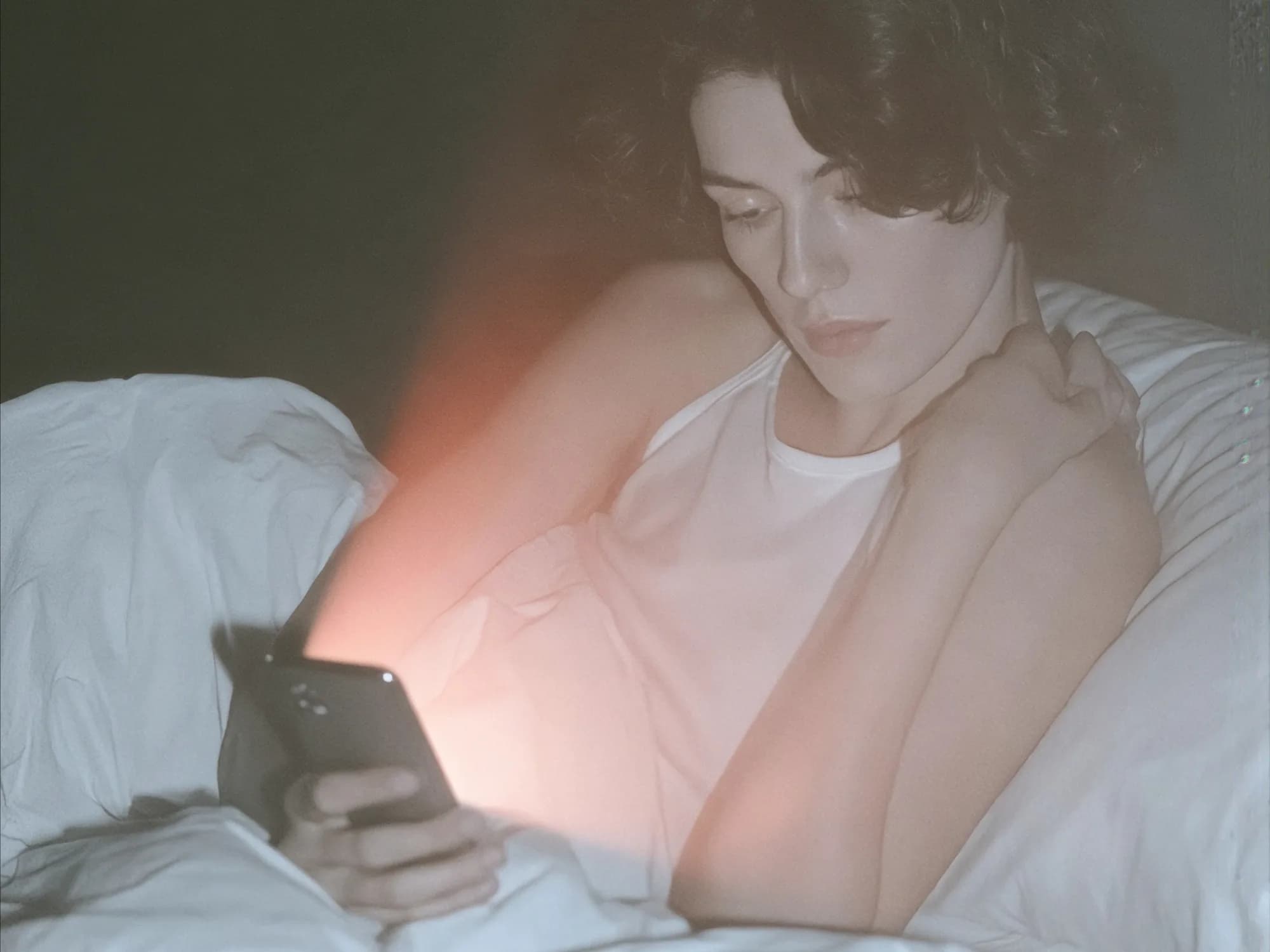 A woman sits up in bed at night, looking at her phone illuminated by a warm glow.