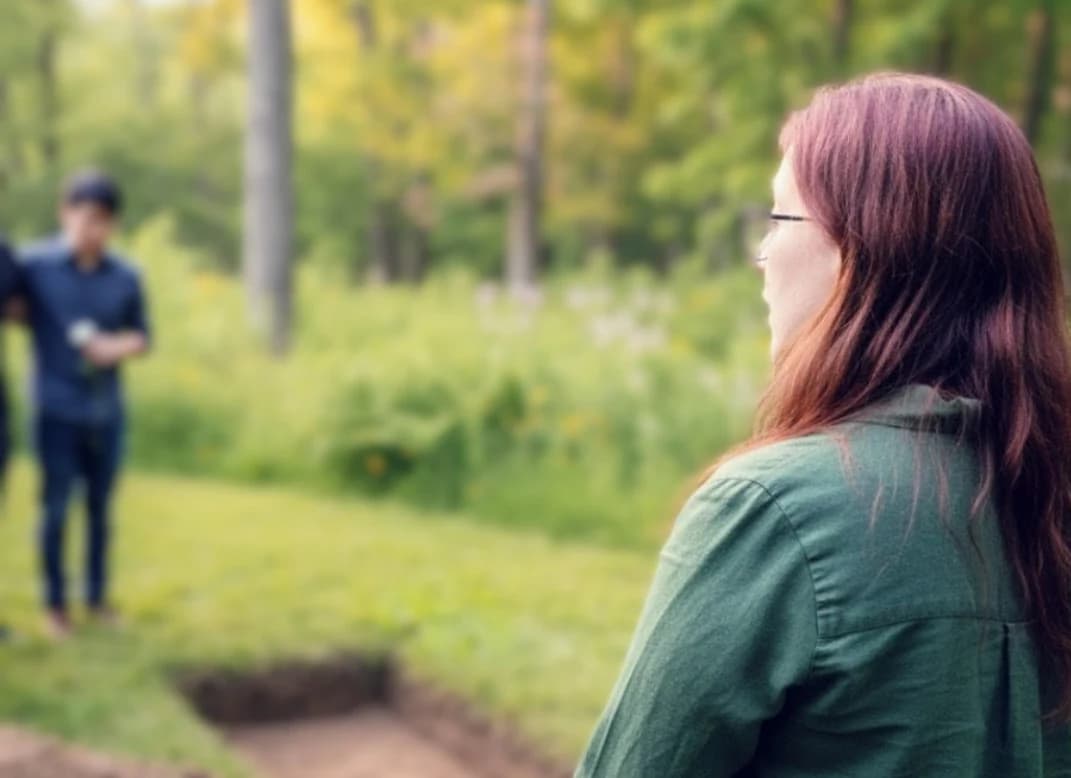 Elayne Sallemi standing at the edge of an open grave in a wildflower meadow during a green burial, with a colleague nearby.