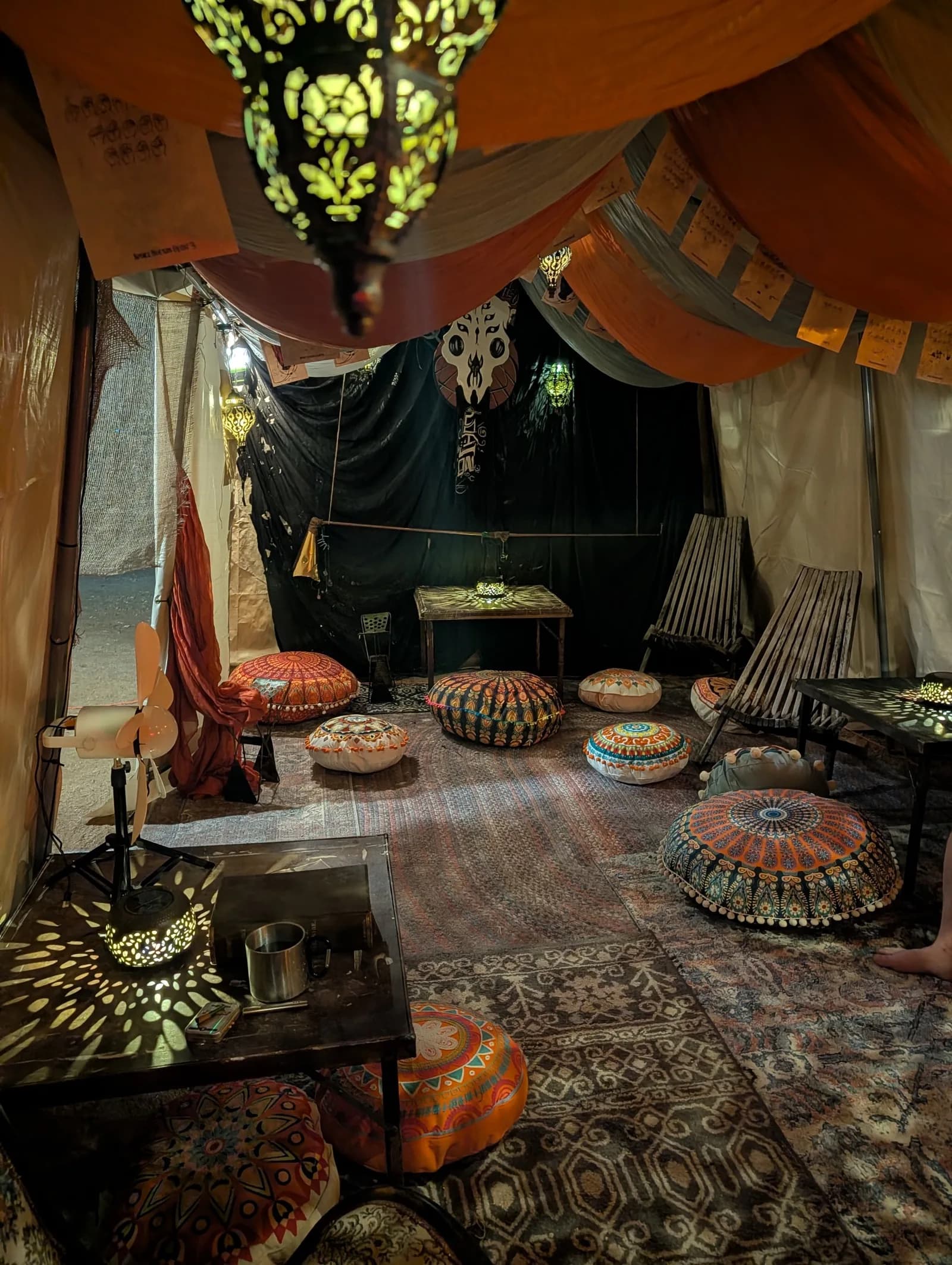 Inside a camp tent draped in orange and grey fabric, lit by pierced-metal lanterns, with mandala floor cushions and patterned rugs.