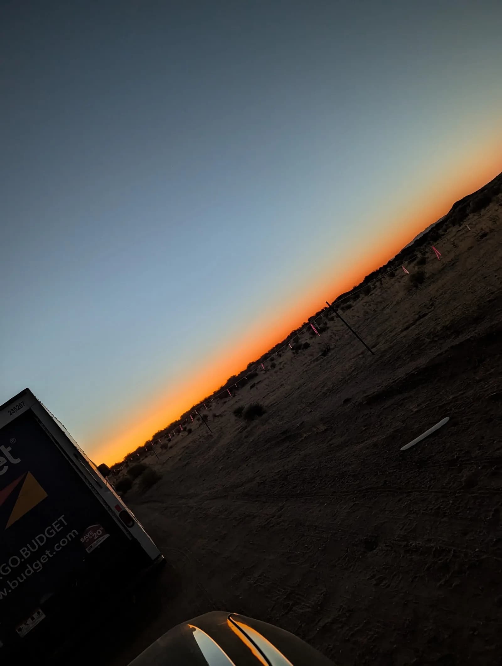 The desert horizon at dusk, a band of orange light fading into deep blue sky above a dirt road and the back of a parked truck.