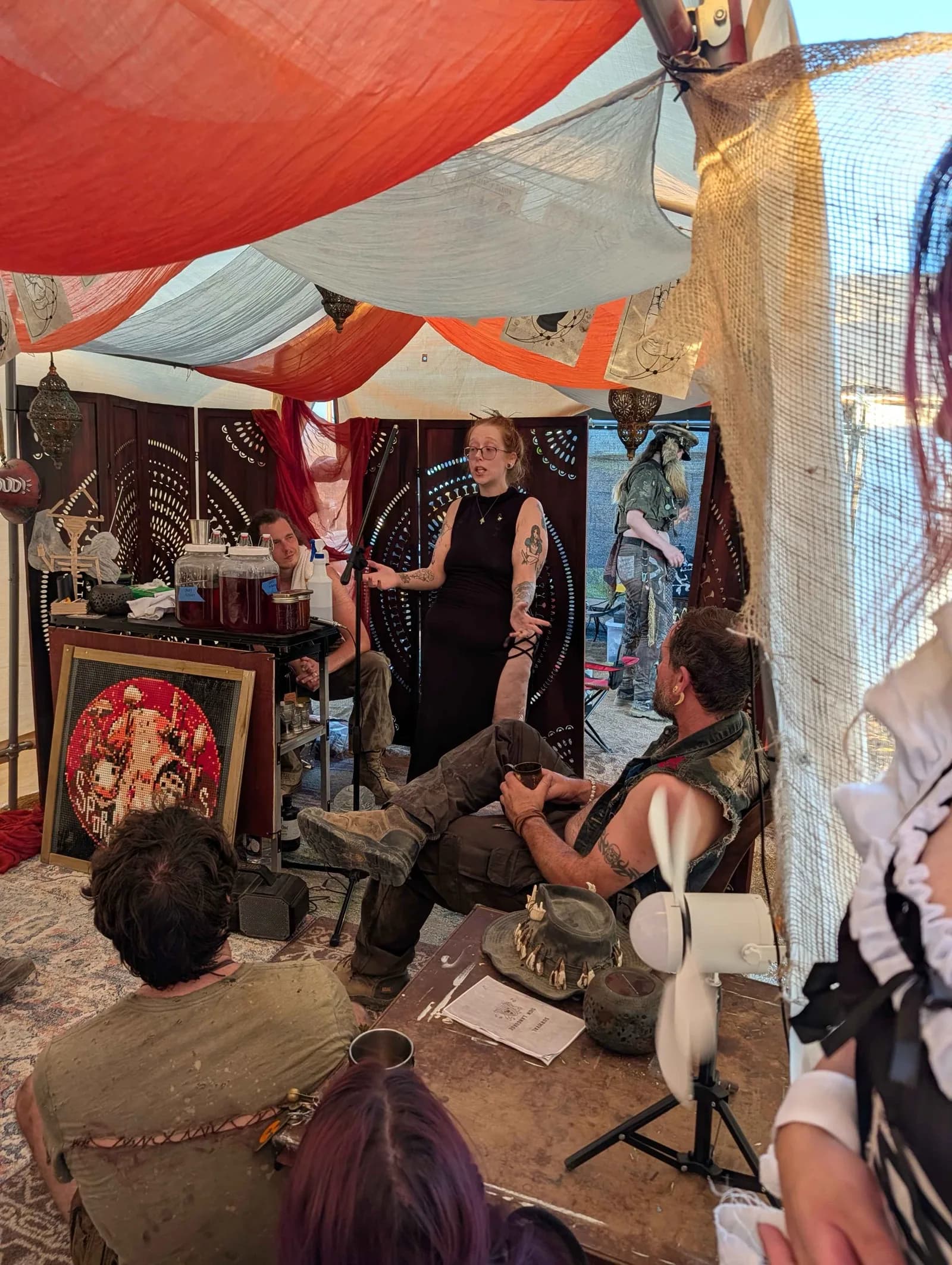 A woman in a black dress stands at a microphone beneath draped fabric, speaking to people gathered in the tent.