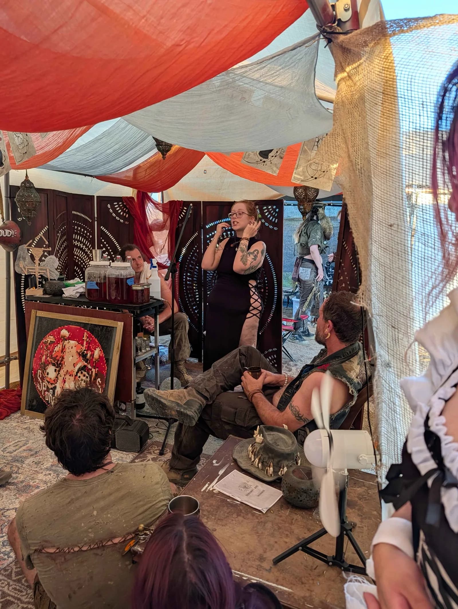 A woman in a black dress speaks with open hands to a group seated around her in the tent.