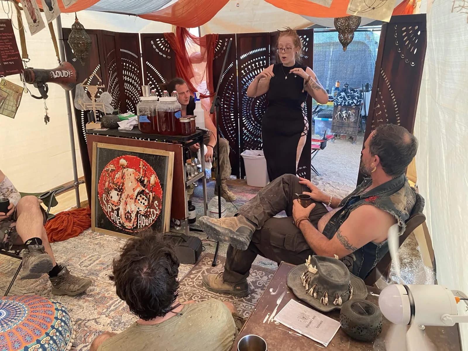 A woman in a black dress speaks to a small group seated in a draped tent, gesturing as she talks.